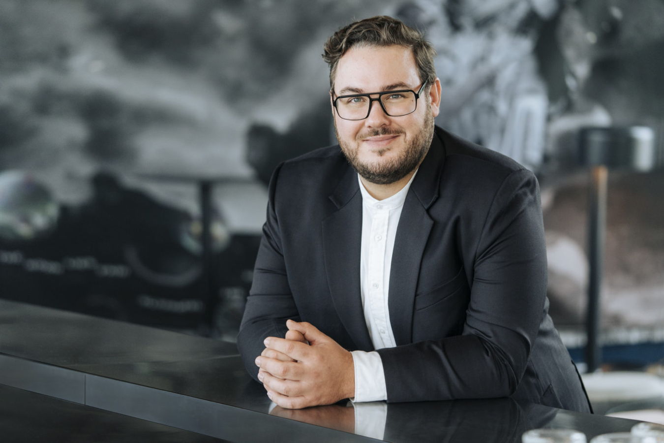 Porträt eines Mannes im schwarzen Anzug und mit Brille, der an einem Tisch sitzt und lächelt