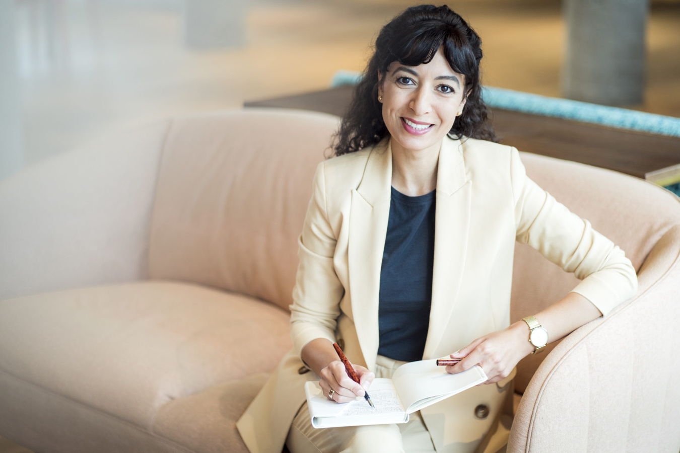 Eine Frau im hellen Blazer sitzt auf einem Sofa und schreibt in ein Notizbuch