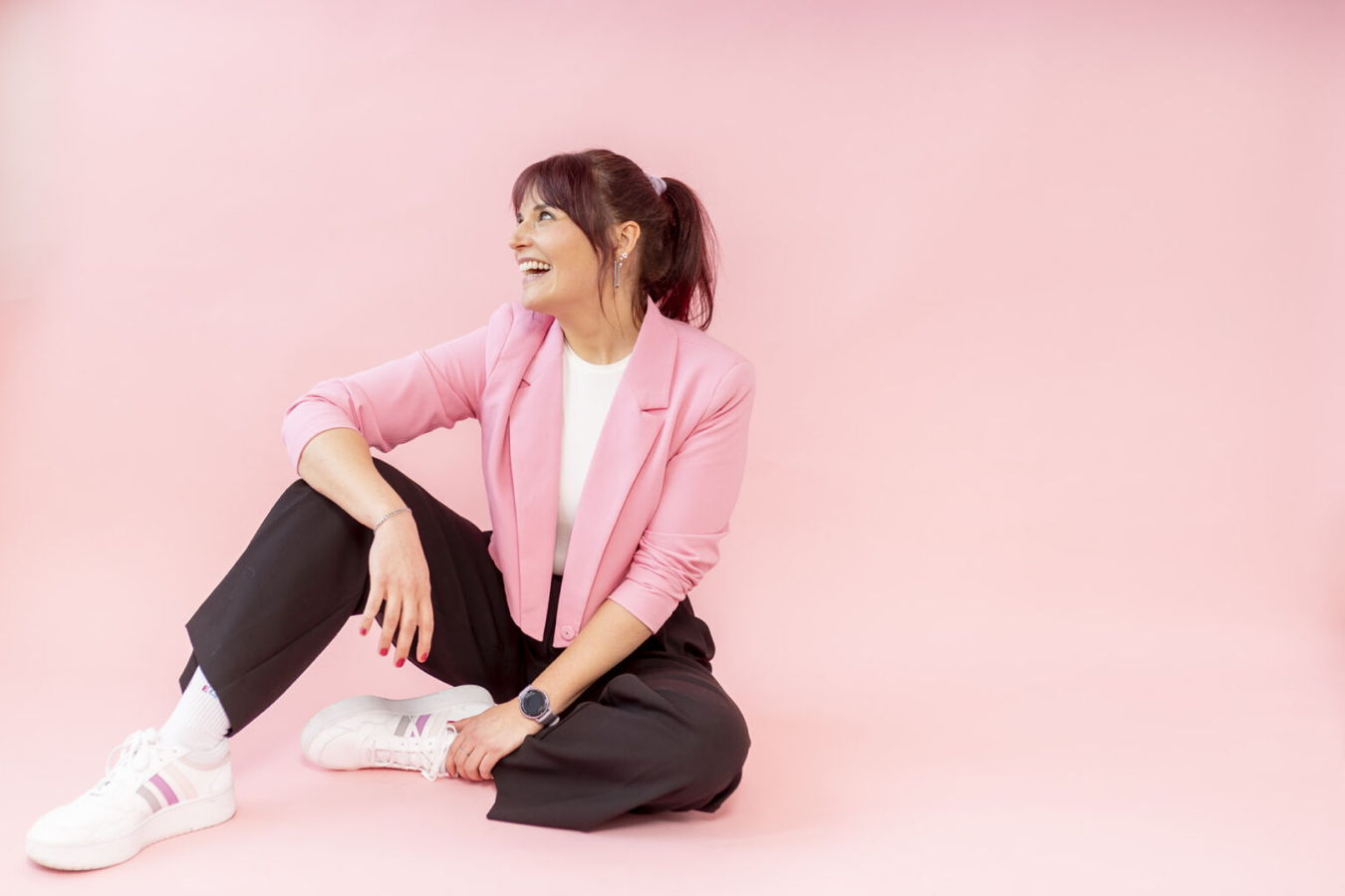 Frau in pinkem Blazer und schwarzer Hose sitzt lächelnd vor rosa Hintergrund