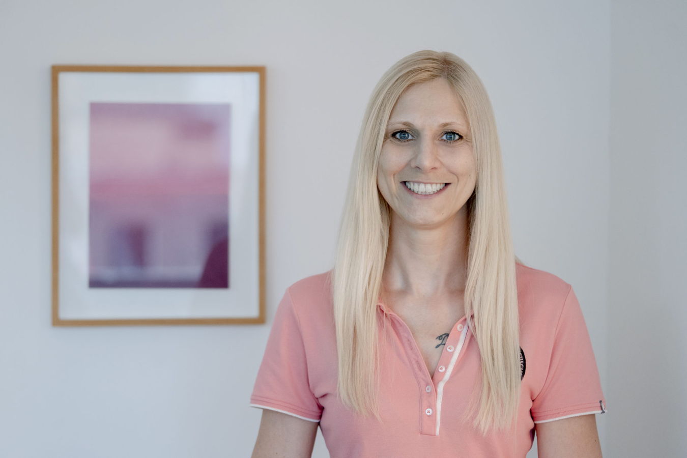 Lächelnde blonde Frau in einem rosa Poloshirt vor einer hellen Wand mit gerahmtem Bild
