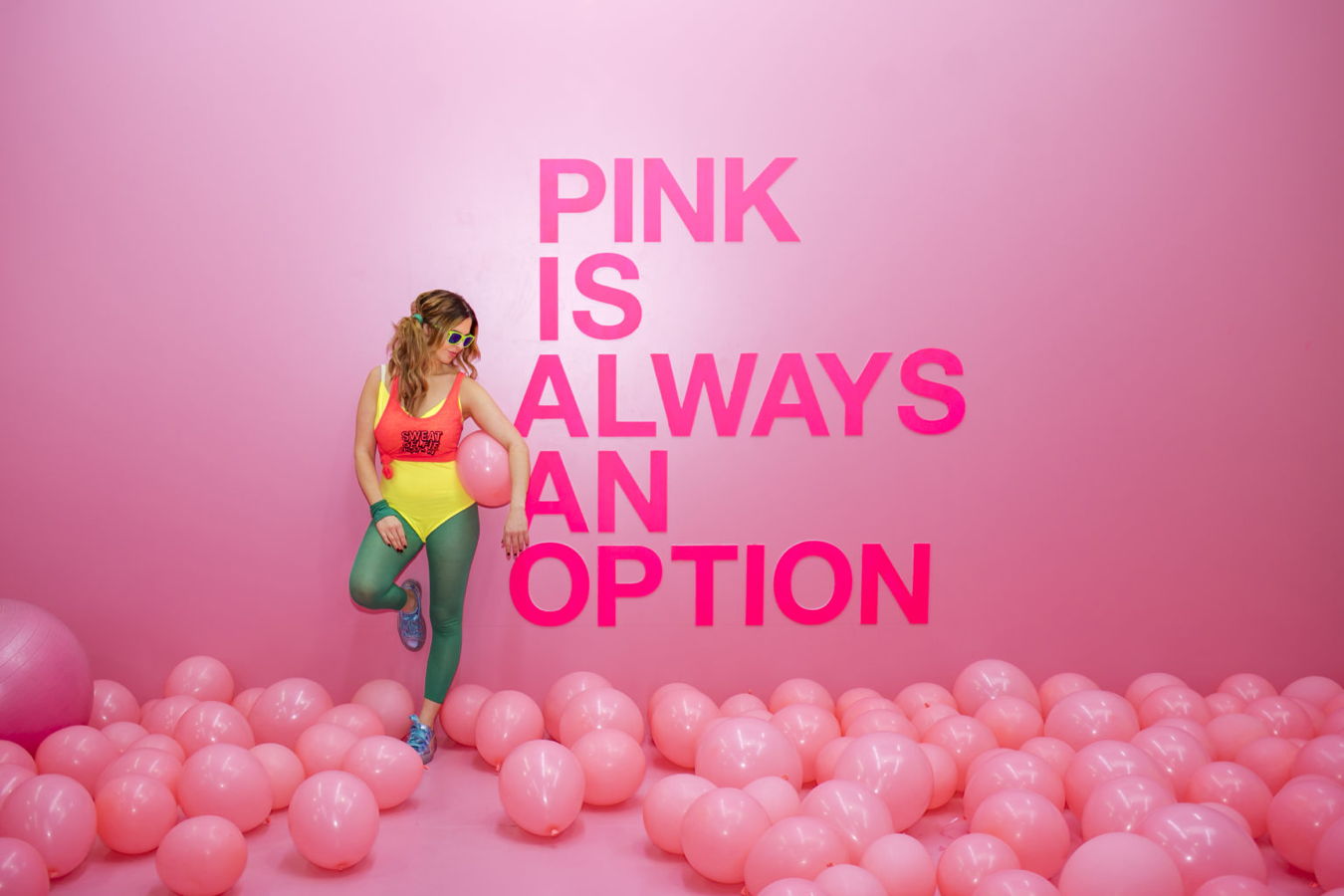 Frau in buntem Outfit vor pinker Wand mit der Aufschrift „PINK IS ALWAYS AN OPTION“ und vielen rosa Luftballons