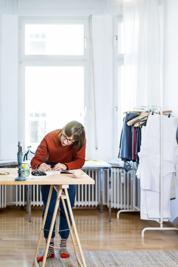 Eine Frau entwirft an einem Tisch in einem hellen Atelier Kleidung