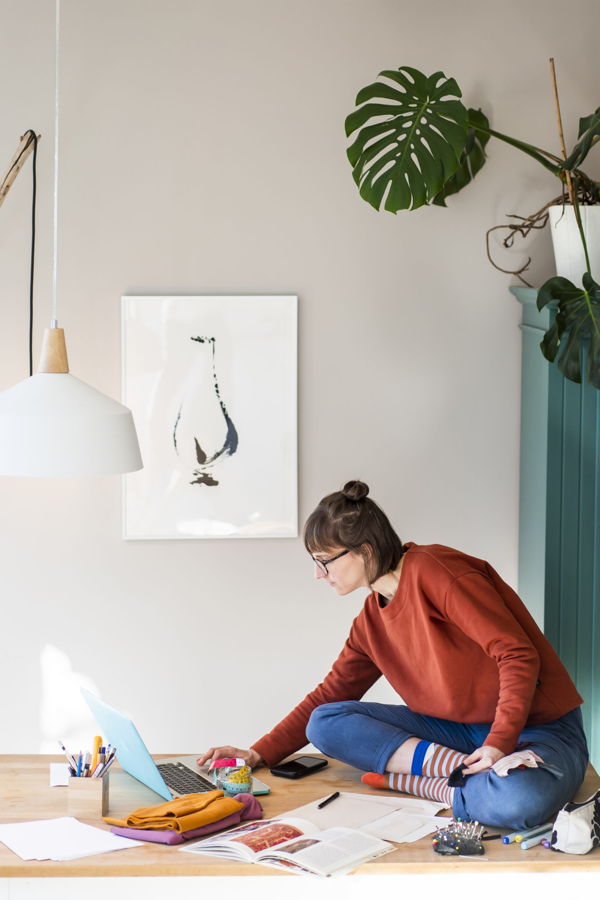 Frau sitzt auf einem Tisch und arbeitet am Laptop, umgeben von Bastelmaterial und Pflanzen