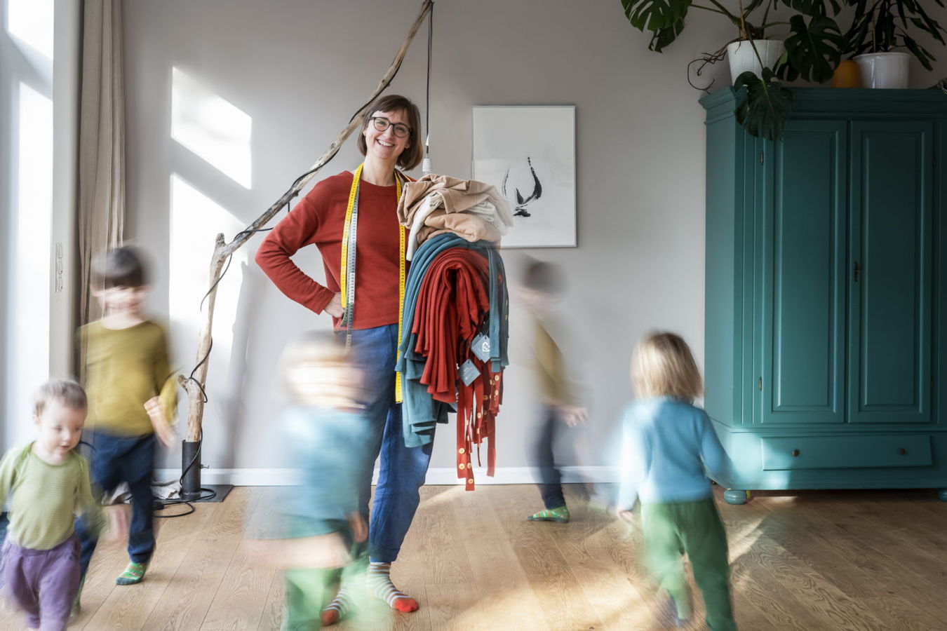 Frau mit Maßband hält einen Stapel Kleidung, während mehrere Kinder um sie herum laufen