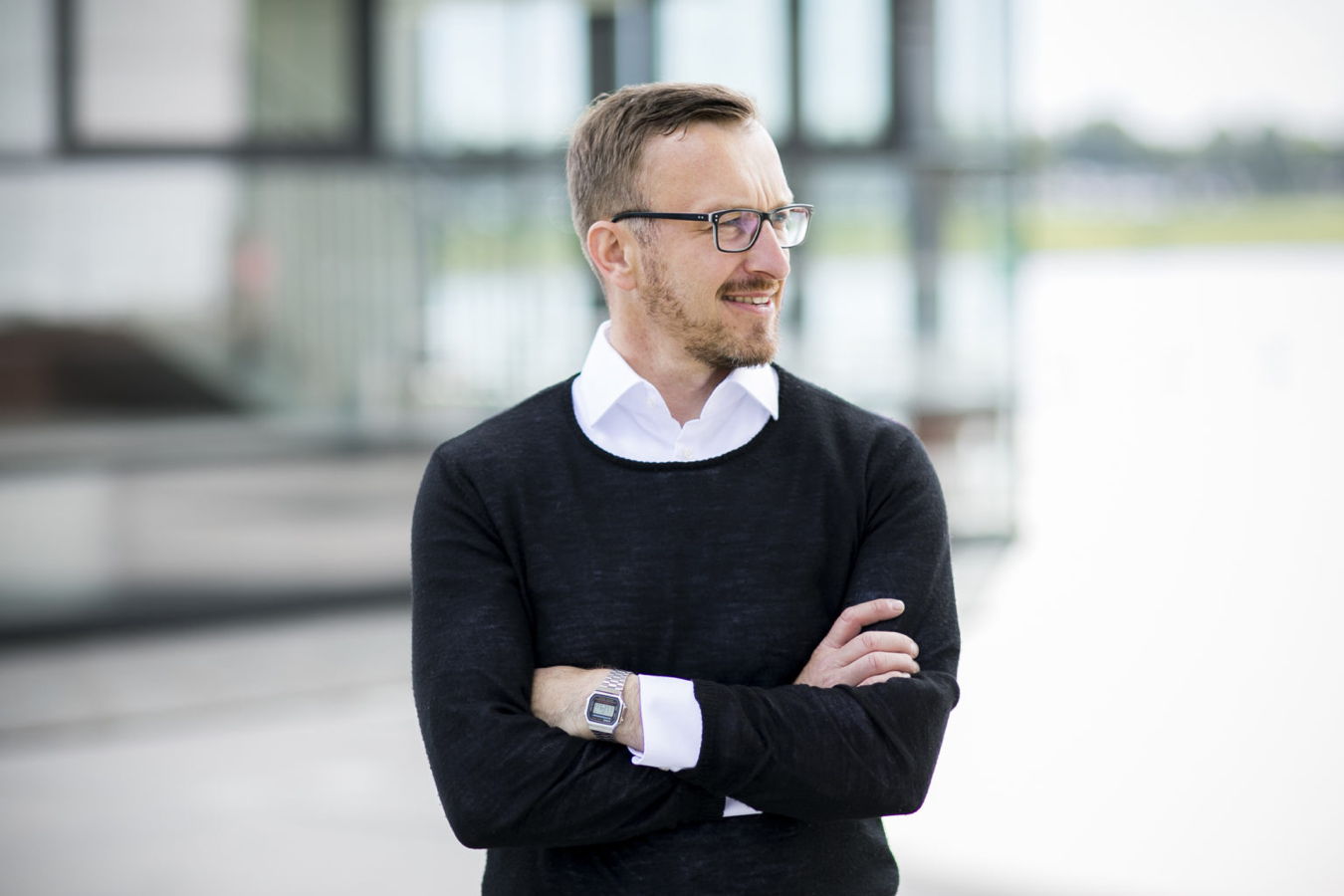 Mann mit Brille und schwarzem Pullover steht mit verschränkten Armen im Freien