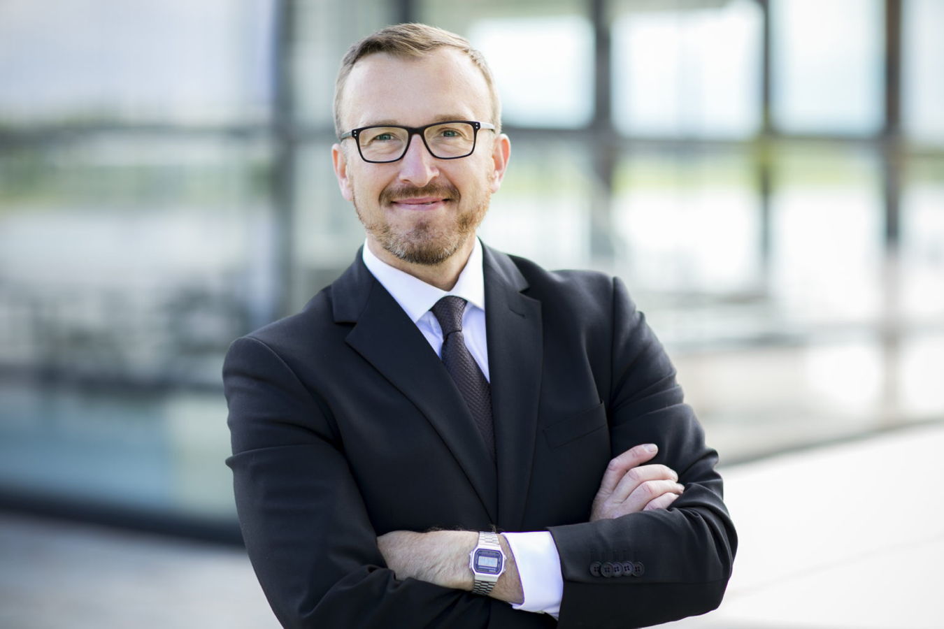 Porträt eines lächelnden Mannes im Anzug mit Brille, die Arme vor der Brust verschränkt