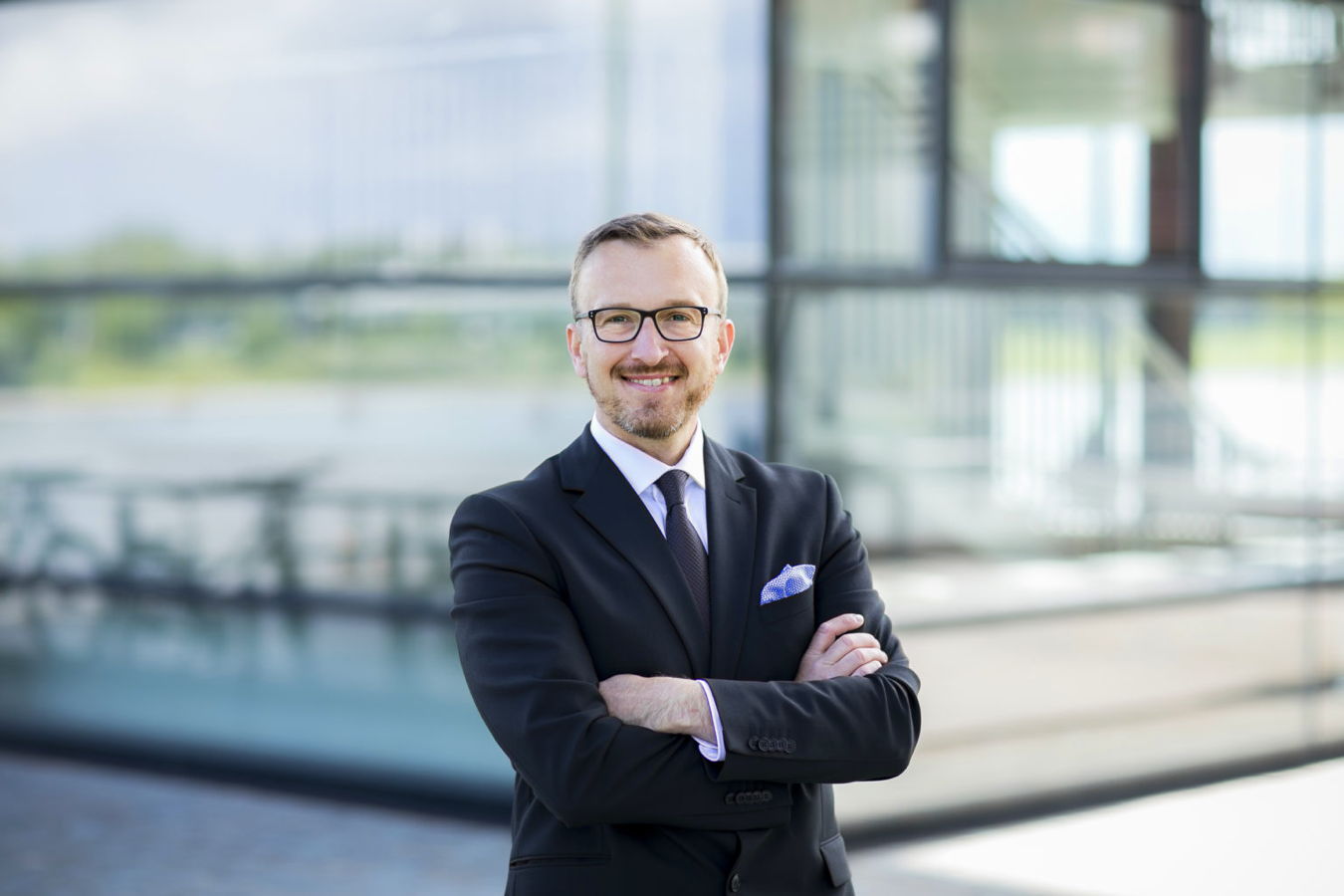 Ein lächelnder Mann im Anzug mit verschränkten Armen vor einem modernen Glasgebäude