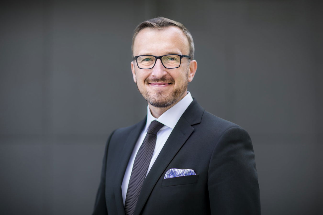 Porträt eines lächelnden Mannes im Anzug mit Brille vor grauem Hintergrund