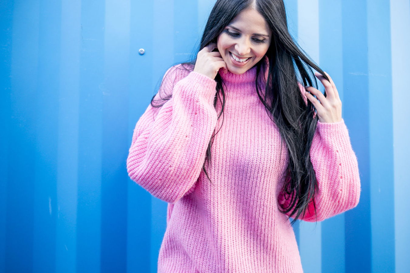 Frau in rosa Pullover vor blauem Hintergrund, lächelnd und sich die Haare haltend