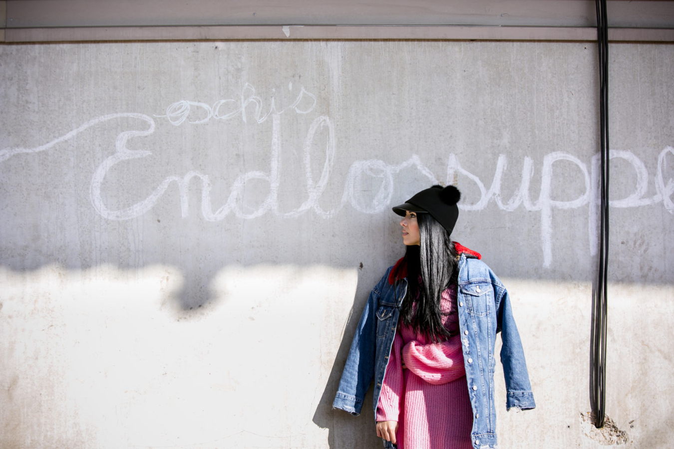 Frau in pinkem Kleid und Jeansjacke vor einer grauen, beschmierten Wand