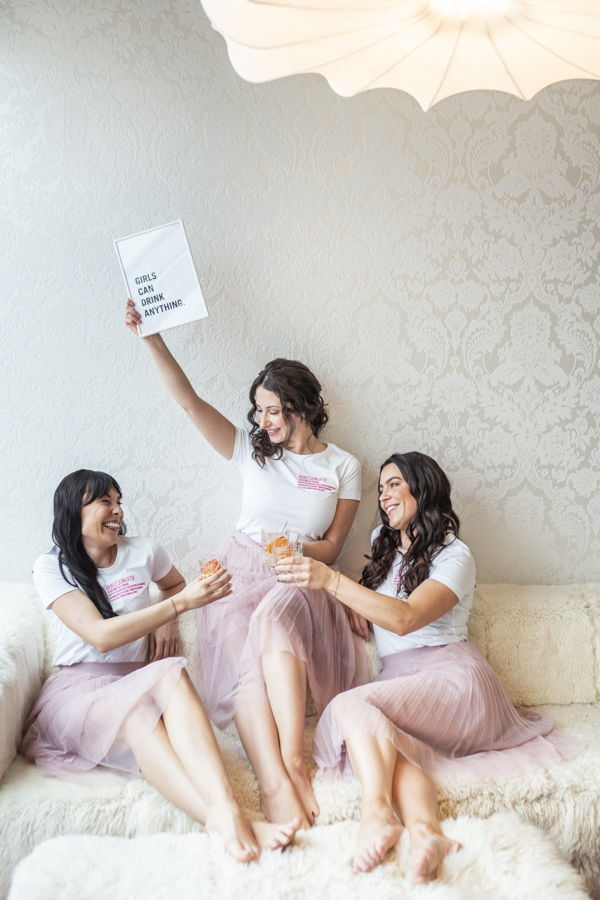 Drei Frauen sitzen auf einem Sofa und stoßen mit Getränken an; eine hält ein Schild mit der Aufschrift „Girls can drink anything.“