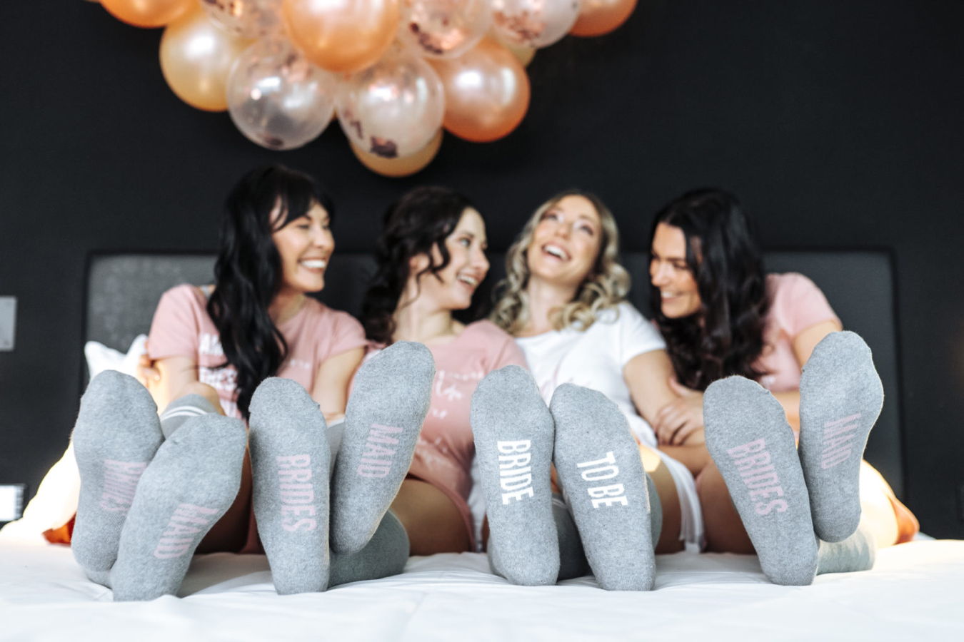 Vier Frauen auf einem Bett mit „Bride“, „To Be“ und „Brides“ auf den Socken, offenbar bei einem Junggesellinnenabschied