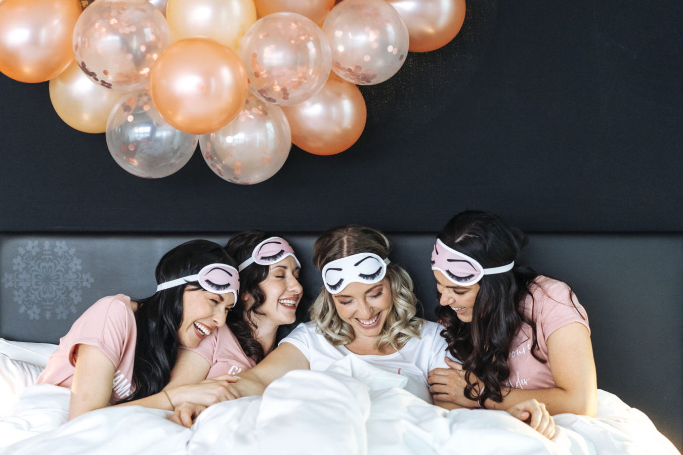 Vier Frauen mit Schlafmasken lachen gemeinsam im Bett unter einer Ballondekoration