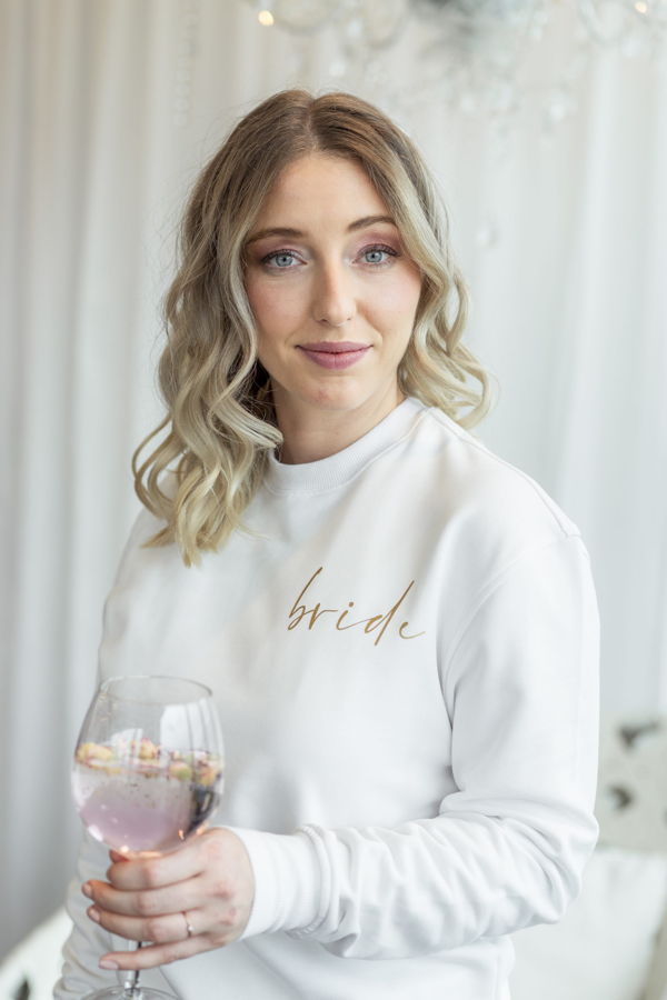 Frau in weißem Sweatshirt mit der Aufschrift „bride“ hält ein Glas mit Getränk