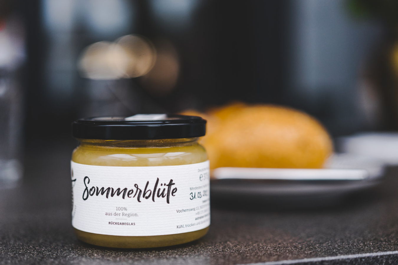 Ein Glas Marmelade mit der Aufschrift „Sommenblüte“ vor einem Teller mit Brot
