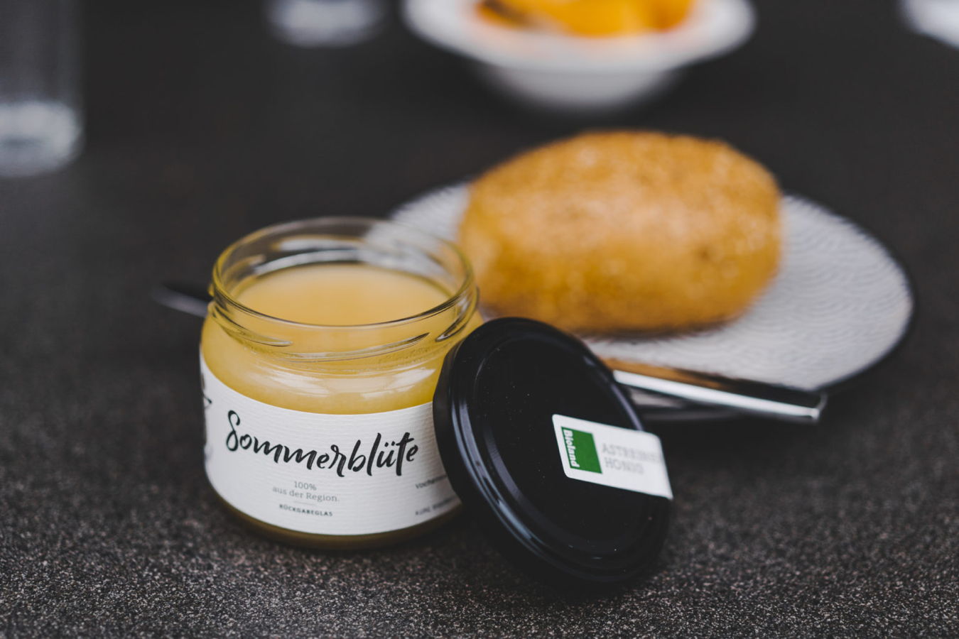 Geöffnetes Glas mit gelb-orangefarbener Creme, daneben Deckel und ein Brötchen auf einem Teller