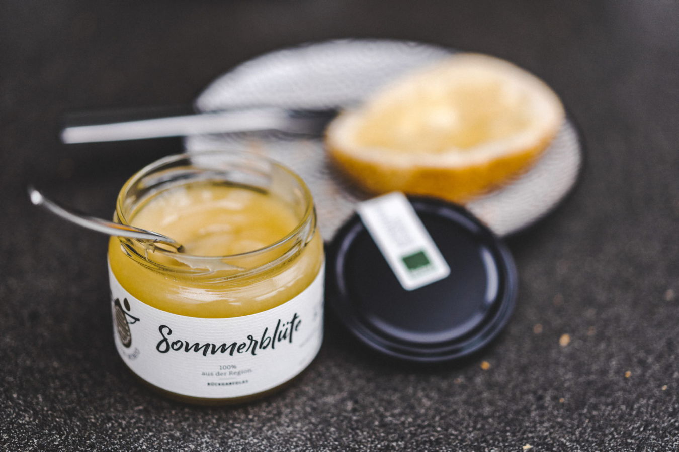 Geöffnetes Glas mit gelbem Aufstrich, Löffel und halbierte Zitrusfrucht daneben