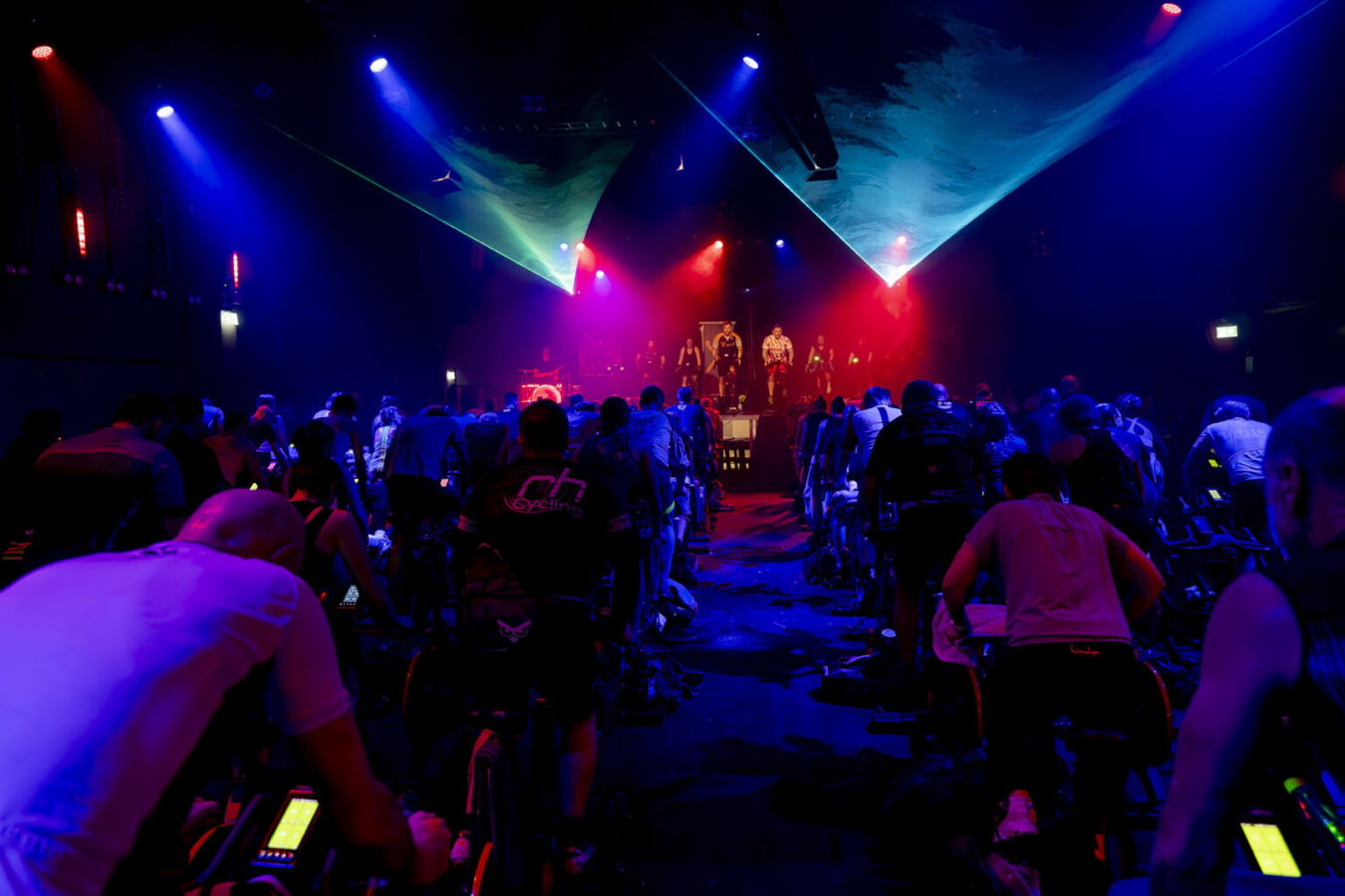 Indoor-Cycling-Kurs mit vielen Teilnehmenden vor einer beleuchteten Bühne