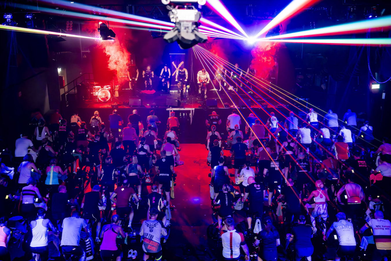 Indoor-Cycling-Kurs mit vielen Teilnehmenden vor einer Bühne unter farbigen Laserlichtern