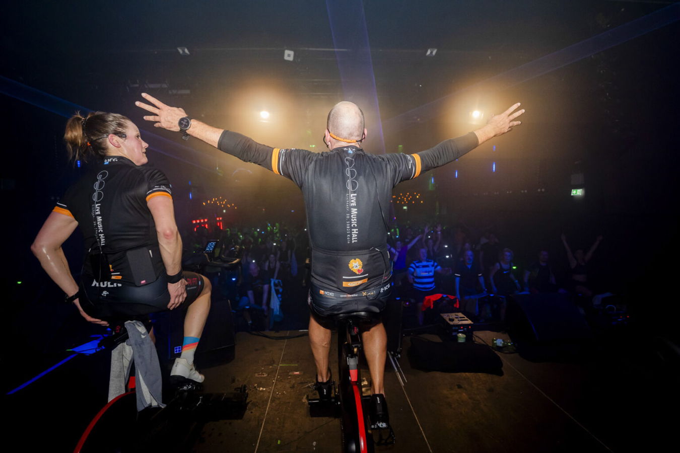 Zwei Indoor-Cycling-Trainer auf der Bühne vor jubelndem Publikum, mit ausgebreiteten Armen unter Scheinwerfern