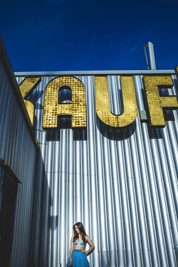 Frau in blauem Outfit vor einer silbernen Metallfassade mit großem gelbem Schriftzug „KAUF“