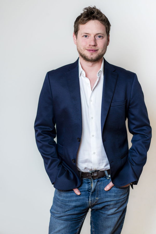 Porträt eines Mannes in blauem Sakko, weißem Hemd und Jeans vor hellem Hintergrund