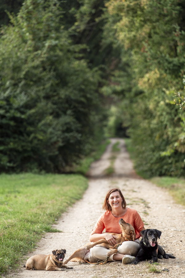 Eine Frau sitzt auf einem Waldweg und umarmt drei Hunde