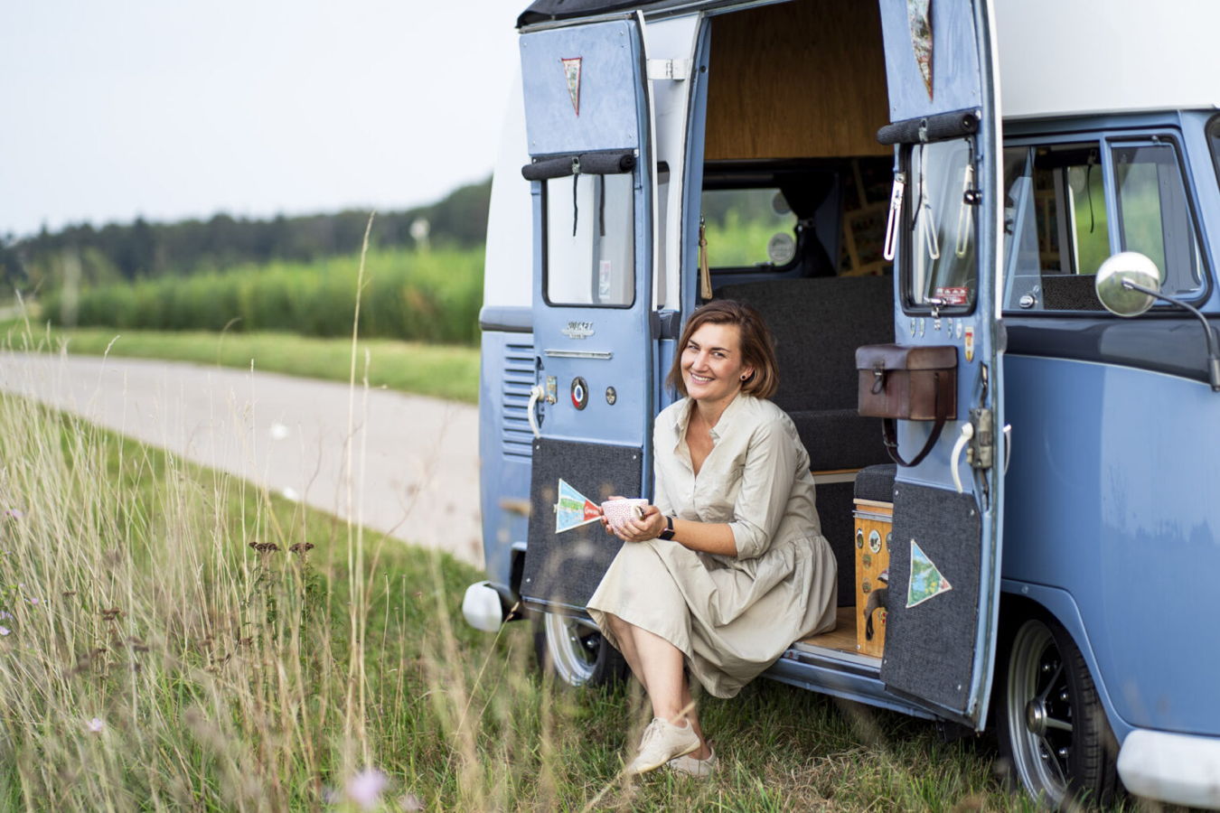 Frau sitzt lächelnd in einem blauen Oldtimer-Wohnmobil mit geöffneten Türen am Feldrand