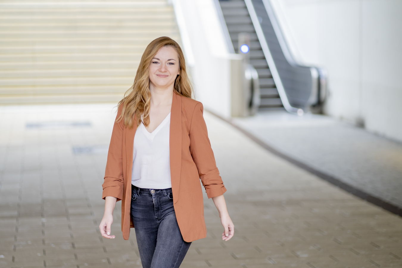 Eine Frau in orangefarbener Jacke geht in einer hellen Passage auf die Kamera zu