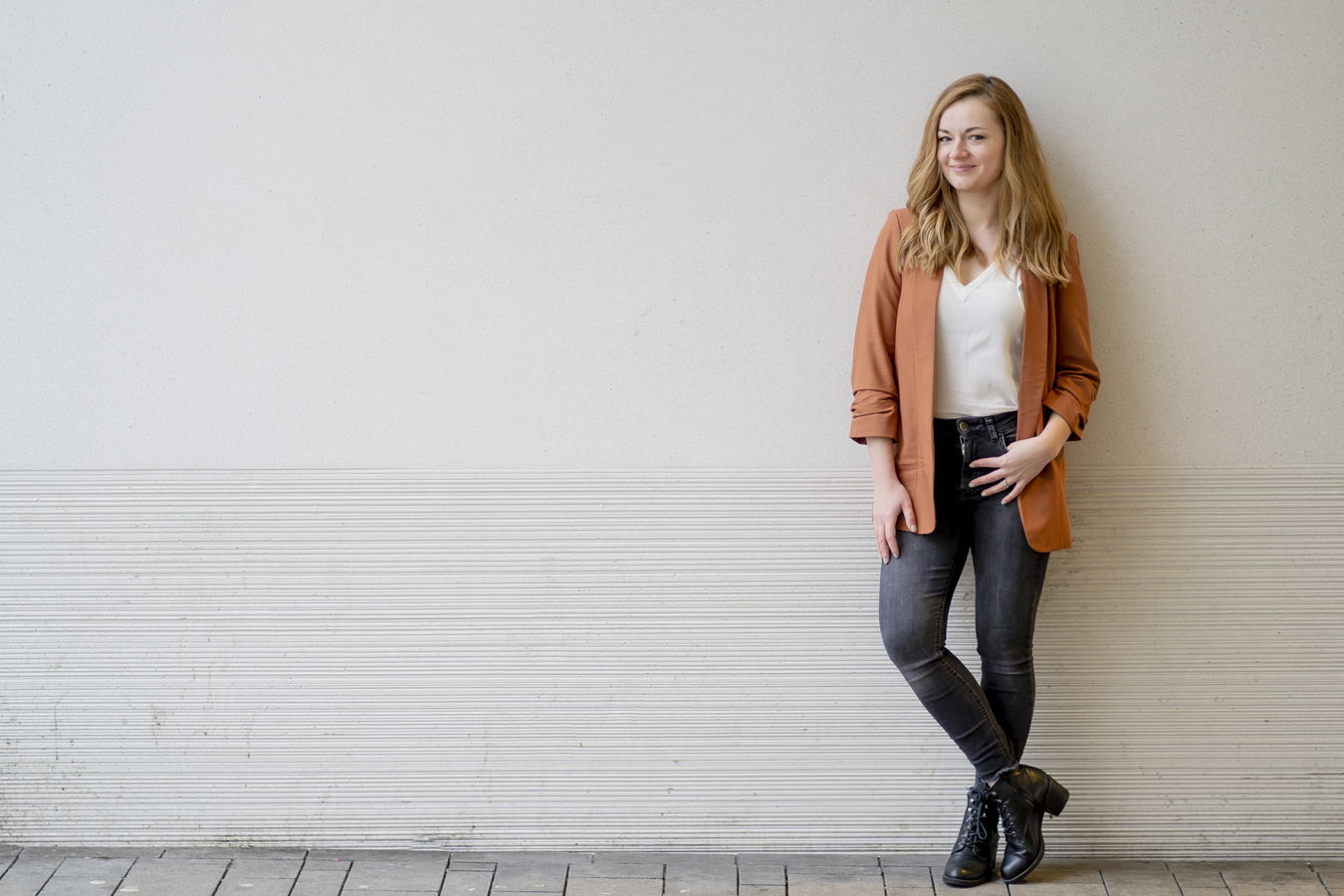 Frau lehnt lässig an einer Wand und trägt orangefarbenen Blazer, weißes Oberteil, Jeans und schwarze Stiefel
