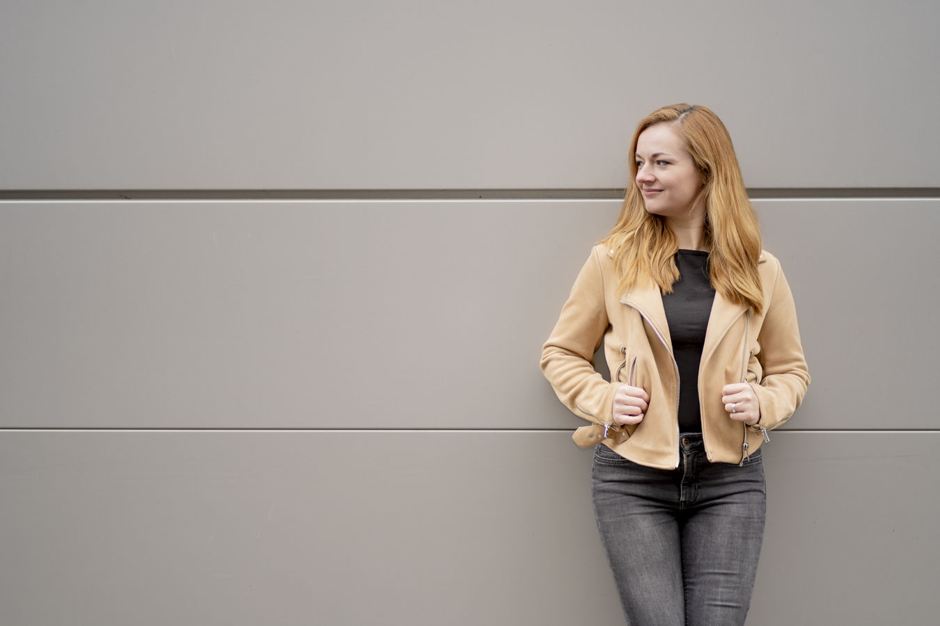 Frau mit blondem Haar in beiger Jacke und Jeans steht vor einer grauen Wand und blickt zur Seite