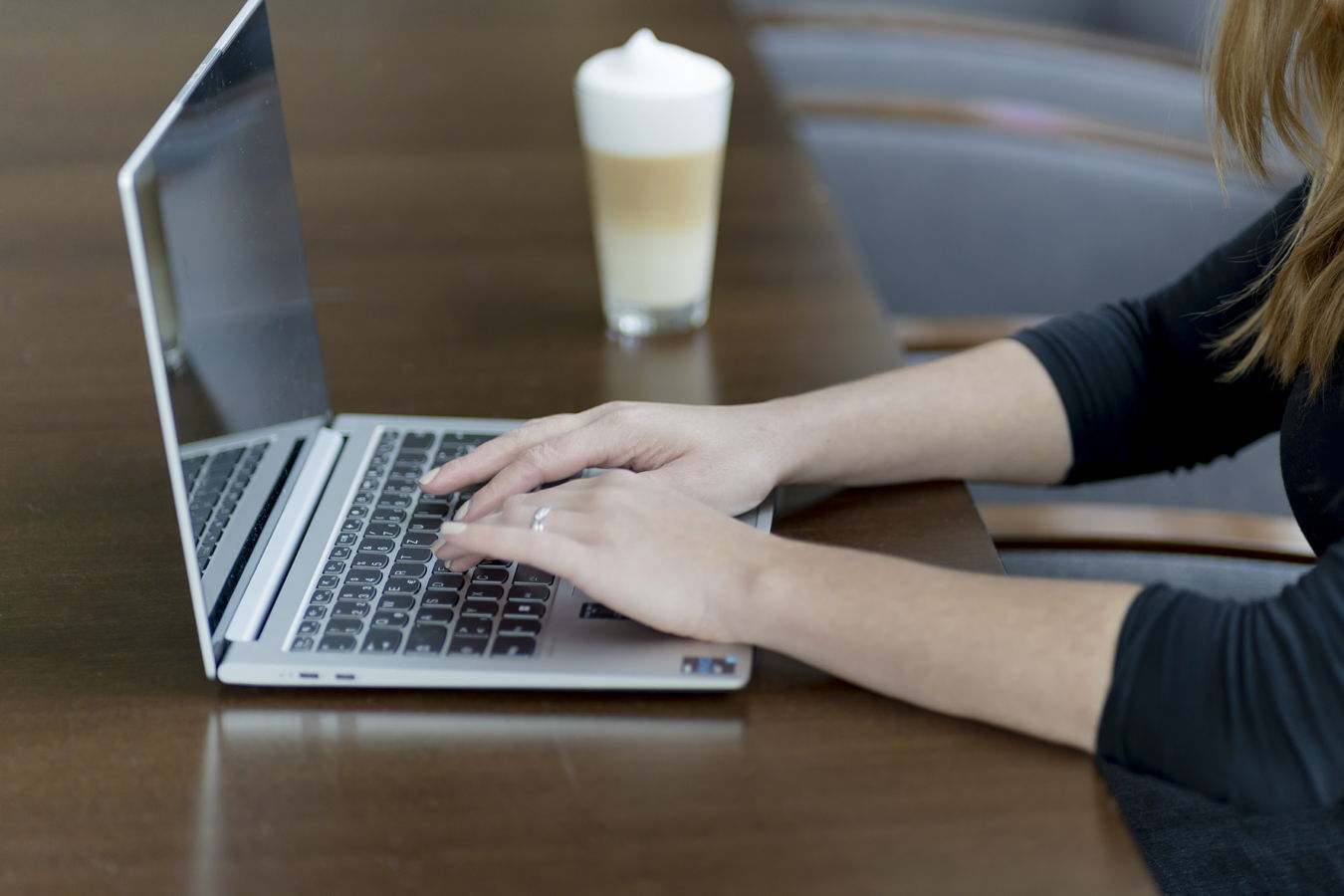 Eine Person tippt an einem Laptop auf einem Tisch; im Hintergrund steht ein Getränk