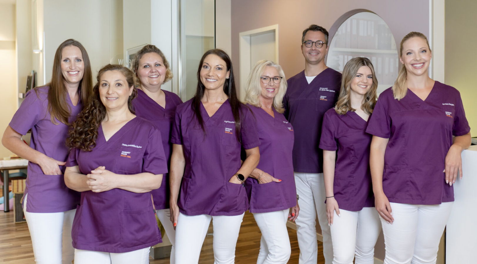 Gruppenfoto von sieben Mitarbeitenden in violetten Praxis-Uniformen in einer Klinik oder Zahnarztpraxis