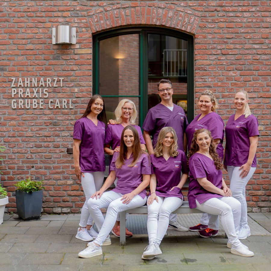 Team der Zahnarztpraxis Grube-Carl vor dem Praxisgebäude in lila Arbeitskleidung