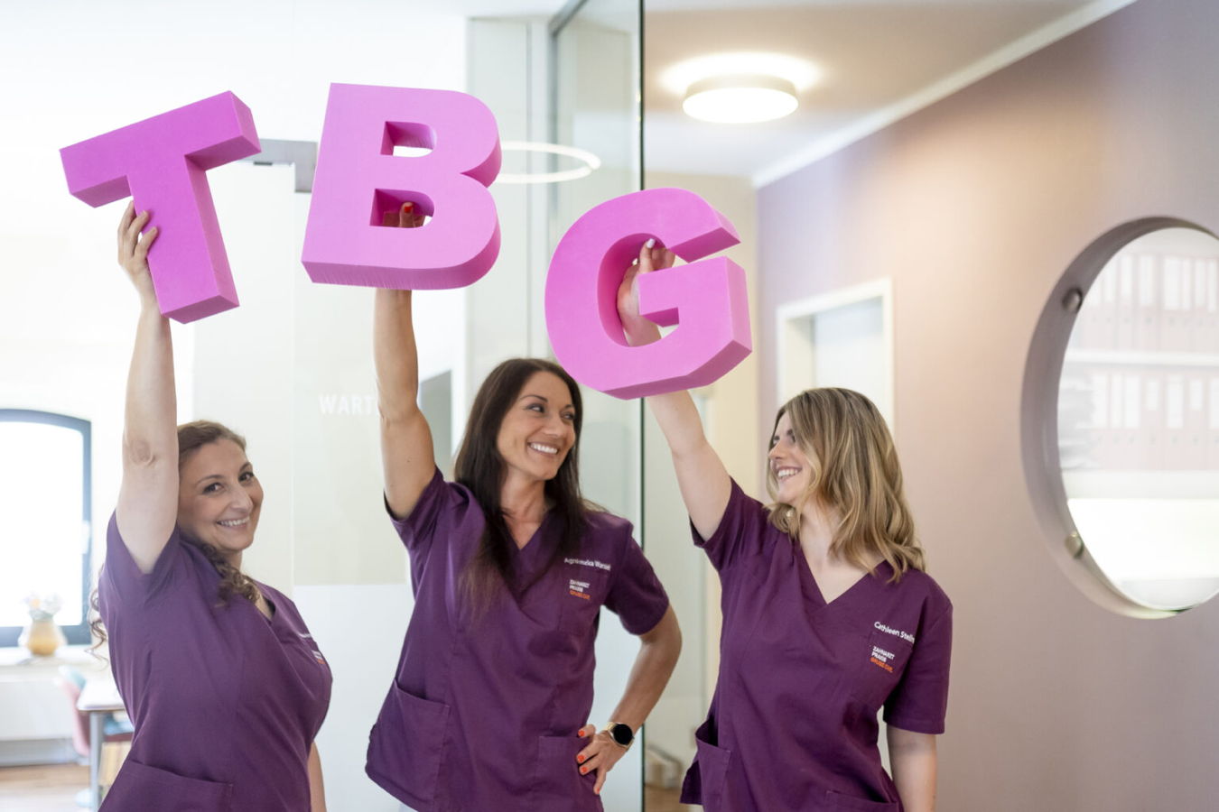 Drei Frauen in violetten Arbeitskleidern halten große pinke Buchstaben T, B und G hoch