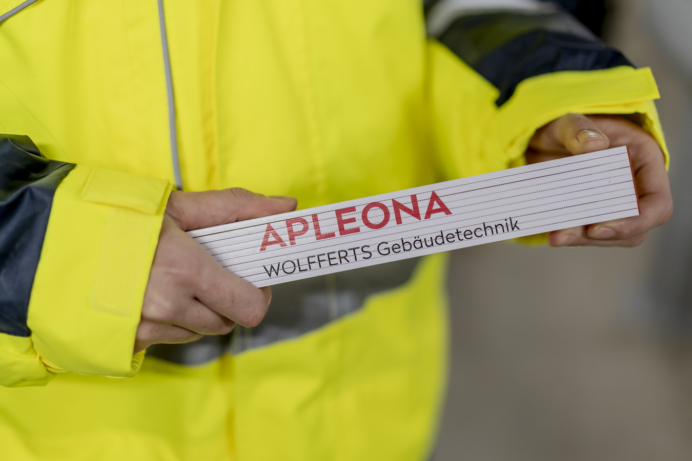 Person in gelber Warnjacke hält ein Schild mit der Aufschrift „APLEONA WOLFFERTS Gebäudetechnik“