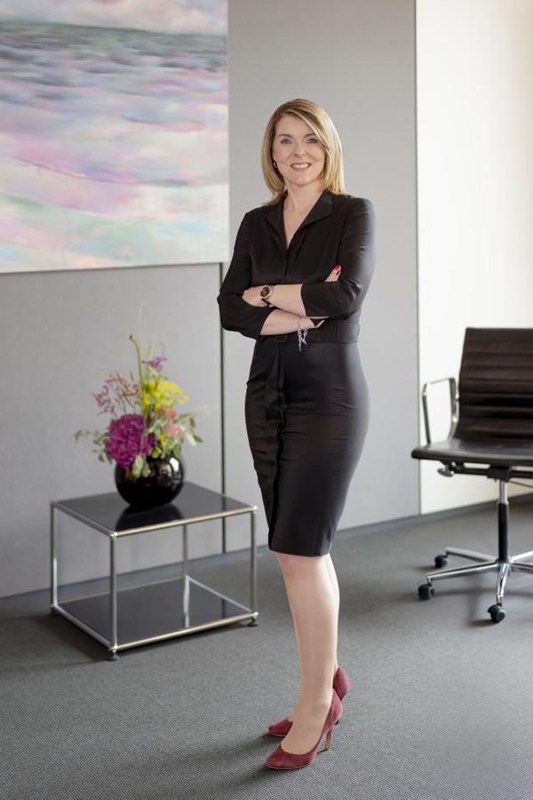 Frau in schwarzem Business-Kleid steht mit verschränkten Armen in einem modernen Büro