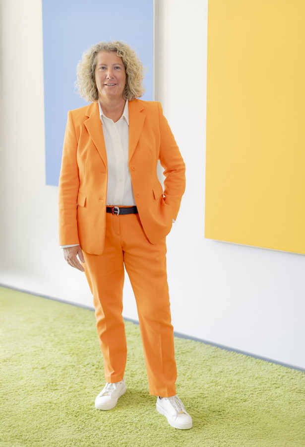 Frau in einem orangefarbenen Anzug steht in einem hellen Raum vor farbigen Wandflächen