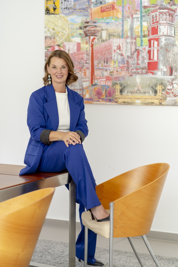 Eine Frau im blauen Anzug sitzt auf einem Tisch in einem hellen Raum vor einem farbigen Gemälde