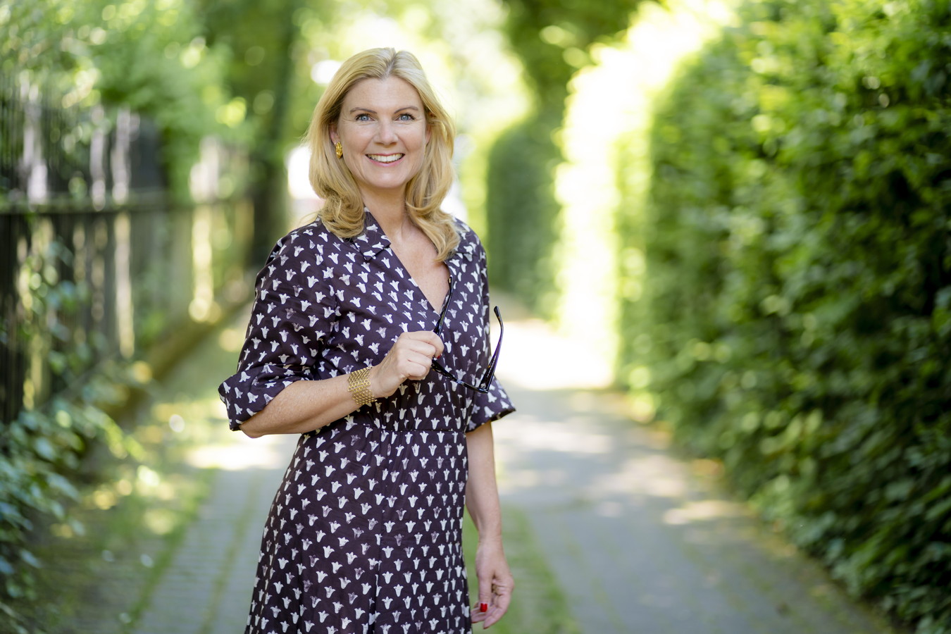 Lächelnde Frau im gemusterten Kleid steht auf einem grünen Weg und hält eine Brille in der Hand