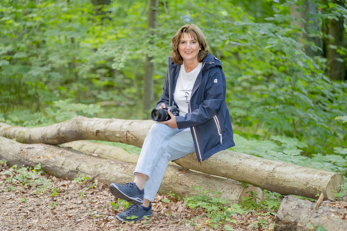 Frau sitzt mit einer Kamera auf einem Baumstamm im Wald