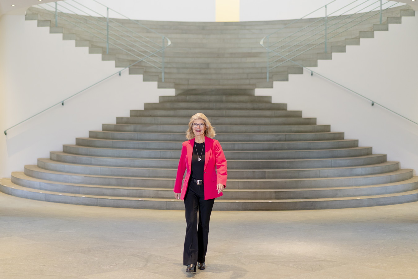 Frau in pinkem Blazer steht vor einer großen geschwungenen Treppe in einem hellen Innenraum