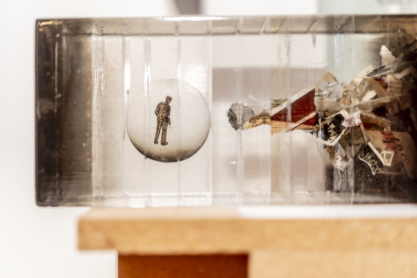 Kleines Figurenobjekt in einer transparenten Box mit Papier- und Materialfragmenten rechts