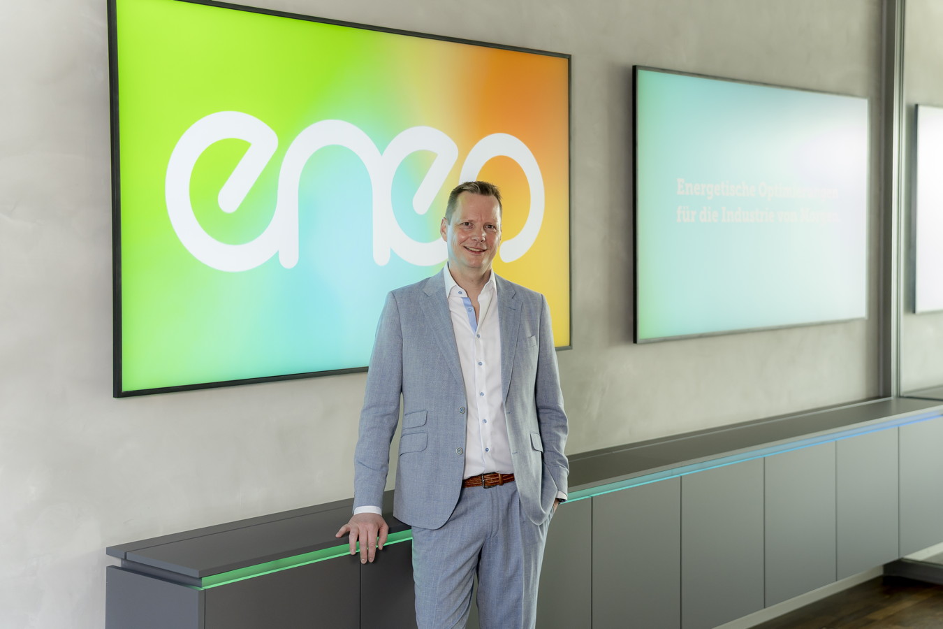 Mann im hellgrauen Anzug steht vor einem farbigen eno-Logo in einer modernen Lobby