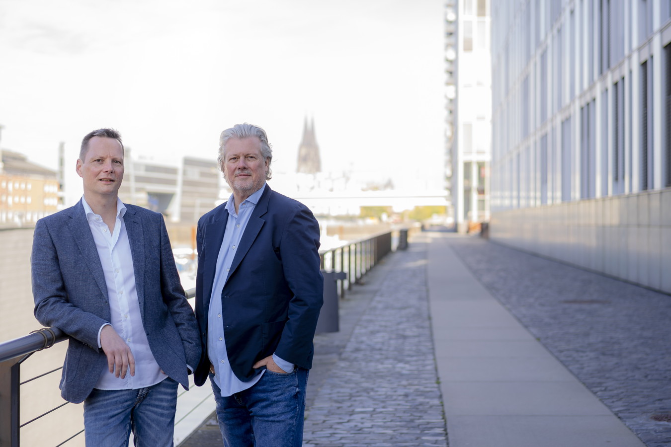 Zwei Männer in Blazern stehen an einer Uferpromenade vor modernen Gebäuden