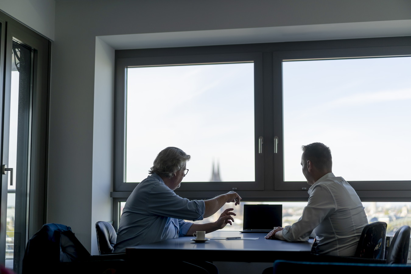 Zwei Männer besprechen sich an einem Tisch vor einem Laptop in einem Büro mit großen Fenstern