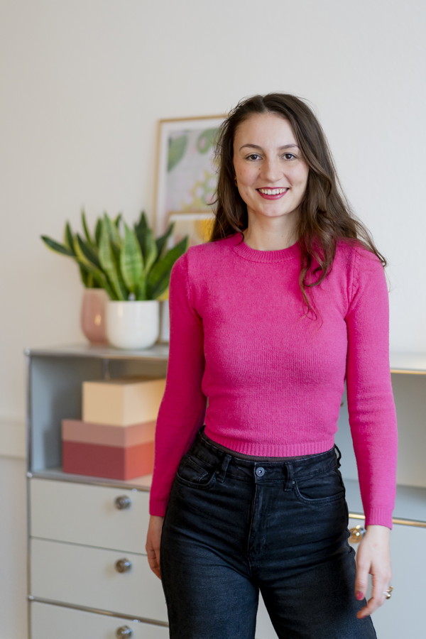 Frau in pinkem Pullover und dunkler Jeans steht in einem hellen Zimmer und lächelt in die Kamera