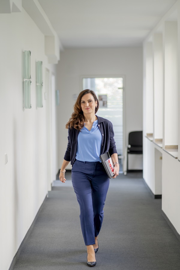 Frau im Business-Outfit geht einen hellen Flur entlang und hält eine Mappe