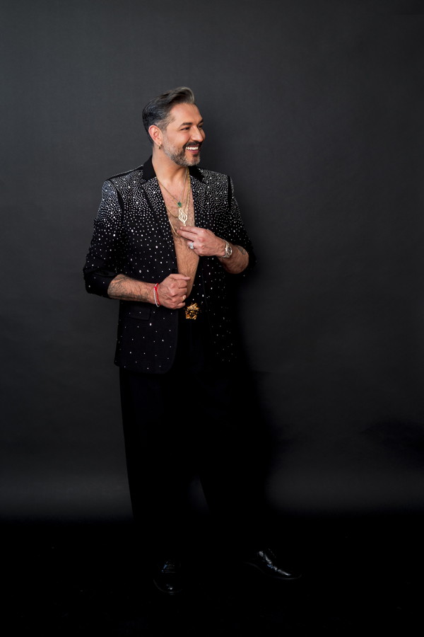 Mann in schwarzem, glitzerndem Anzug vor dunklem Hintergrund, lächelnd zur Seite blickend