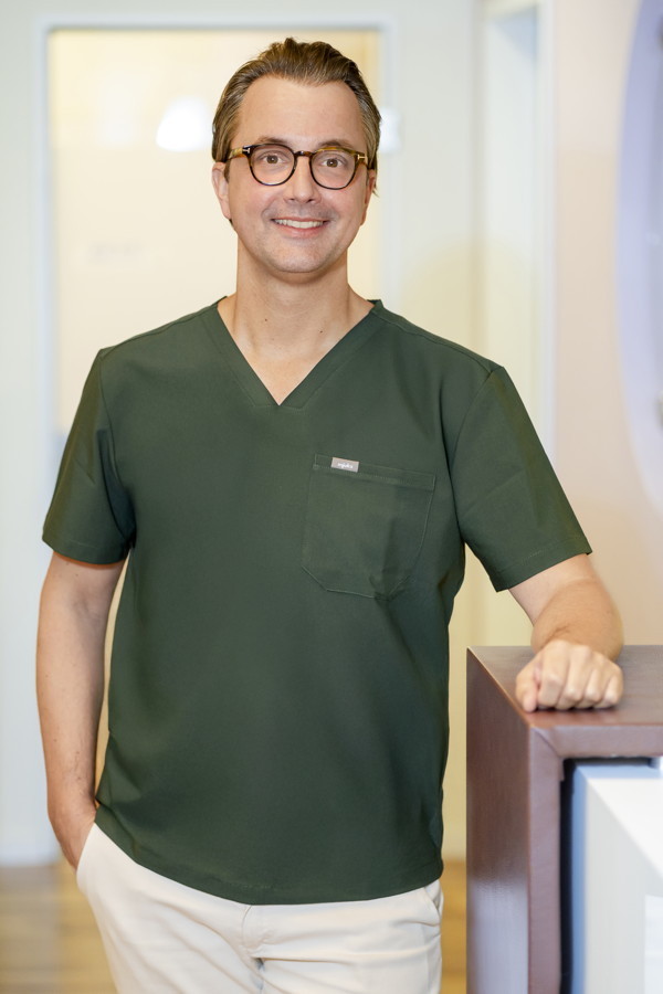 Mann in grüner OP-Kleidung mit Brille steht in einem Klinikflur